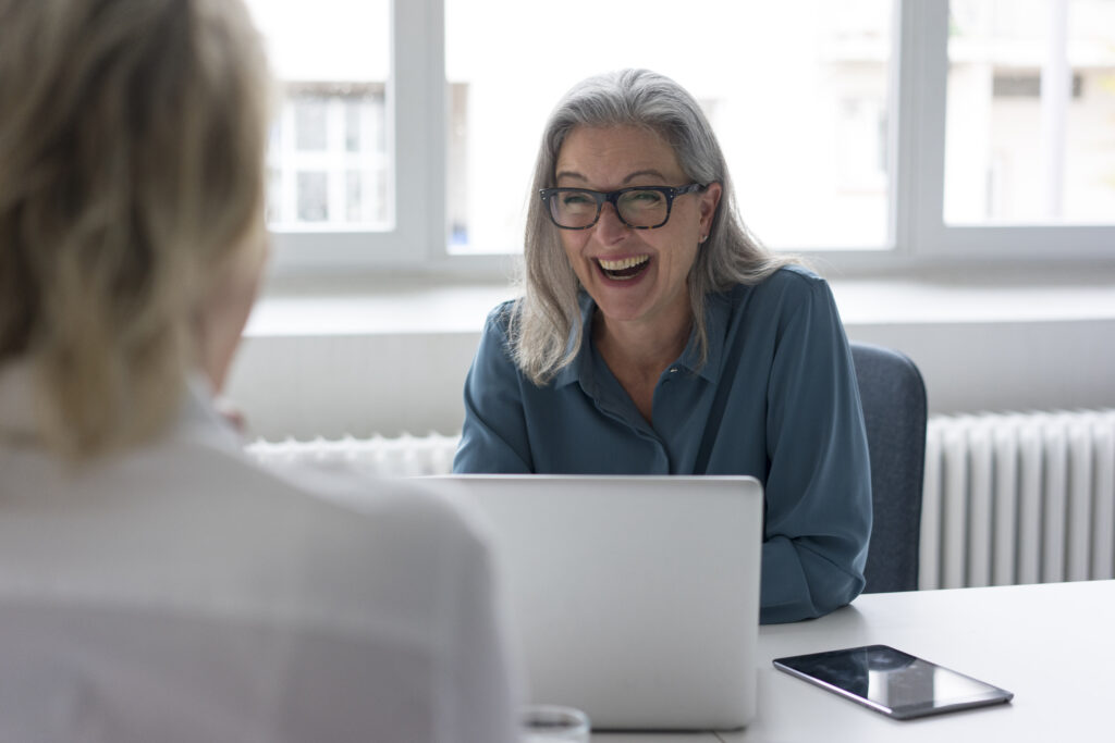 Laughing mature businesswoman talking to young businesswoman at desk in office